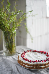A delicious carrot cake stands on the table, garnished with white mascarpone cream and cranberries. A beautiful dessert for every day. There are greens in a vase on the table. Spring still life