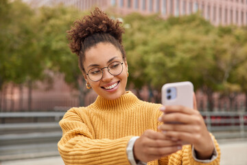 Positive millennial content maker shoots influence video vlog enjoys networking lifestyle takes selfie via smartphone wears round eyeglasses and yellow jumper poses against blurred background - 499669055