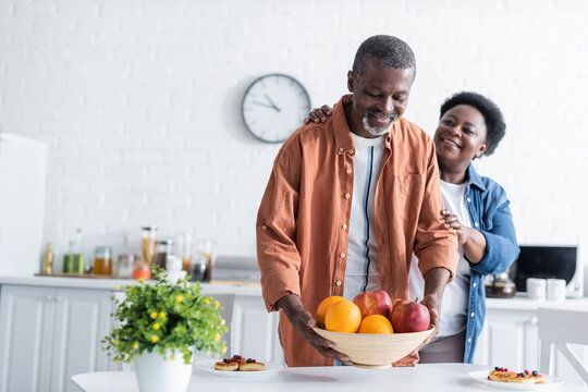 senior african american man putting bowl with fruits on breakfast table near smiling wife.