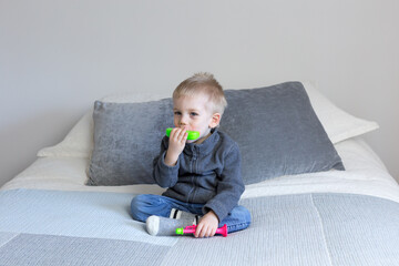 Cute three-year old blond boy sitting on bed dressed in casual clothes playing with a colorful toy harmonica