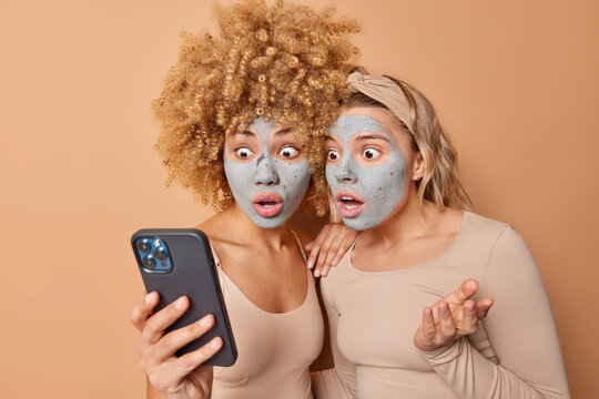 Frightend two women strare scared at smartphone find out shocking news apply beauty masks on face for skin treatment dressed in casual clothes take care of appearance isolated over beige wall