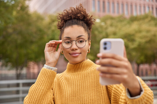 Pleased confident woman with curly hair takes selfie pictures or shoots video for sharing to web blog keeps hand on rim of spectacles dressed in yellow knitted sweater poses at street outdoors