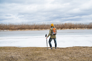 A view from behind a male tourist stands on the edge of the lake looking into the distance at the landscape, a backpack on his back hiking with sticks in the mountains, a stop on the river bank.