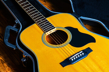 Wooden yellow acoustic guitar lying in a hard case. Stringed musical instrument in a black open wardrobe trunk. Part of the body of a acoustic guitar with fretboard, sound hole and strings close up. - obrazy, fototapety, plakaty
