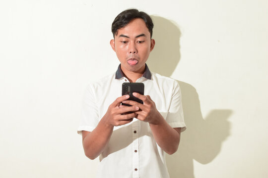 Portrait Of Asian Man Communicating Video Call Using Smartphone. Indonesian Man Posing With His Tongue Sticking Out While Staring At The Cellphone Screen, A Sign Of Mocking Or Joking