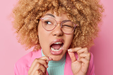 Headshot of young woman with curly hair uses dental floss winks eye and takes care of oral hygiene...