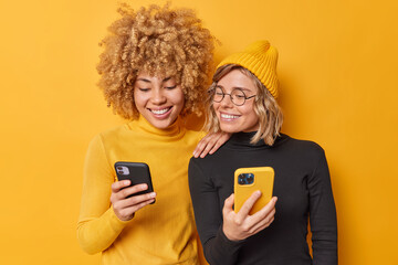 Beautiful two female friends use mobile phones browse internet type sms message dressed in casual turtlenecks smile gladfully isolated over vivid yellow background watch video via media app.