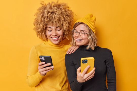 Beautiful two female friends use mobile phones browse internet type sms message dressed in casual turtlenecks smile gladfully isolated over vivid yellow background watch video via media app.