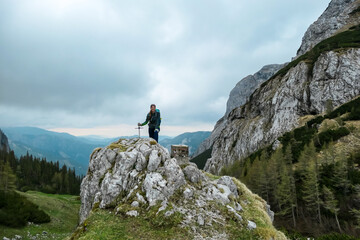 Woman with backpack standing on rock next to the Haeuslalm cottage. Hiking trail in early spring with panoramic view on the mountain peaks of Hochschwab Region in Upper Styria, Austria. Alps, Europe