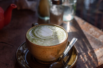 Green Matcha Latte, popular trendy tea with latte art heart shape on top. Served in cafe in ceramic mug on rustic table. Strong shadows and summer sun light