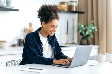 Fototapeta premium Cute successful joyful young african american woman, freelancer, IT specialist, sitting at home in the kitchen, using laptop for work, texting with colleagues, working on a project, smiling happily