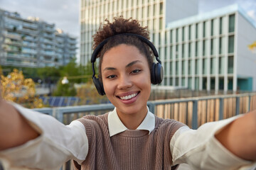 Positive lovely teenage girl with combed curly hair takes selfie in urban setting smiles happily listens music from playlit wears wireless headphones on ears being in good mood during spring day