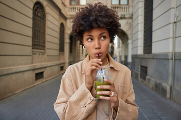 Pensive young woman with curly hair sips fresh vegetable cocktail eats fitness food for weight loss dressed in brown jacket strolls in ancient city thinks about something. Nutrition concept.