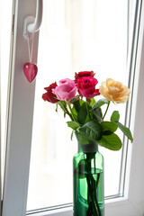 a bouquet of colorful roses in a green vase and a souvenir heart on the background of the window