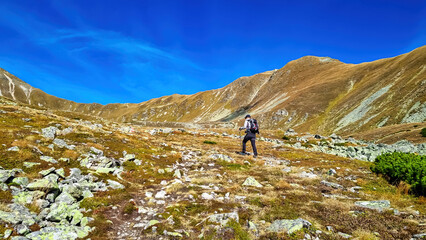 Fototapeta na wymiar Man with backpack on hiking trail leading to Seckauer Zinken in the Lower Tauern mountain range, Styria, Austria, Europe. Sunny golden autumn day in Seckau Alps. Panorama on dry, bare grass terrain