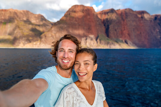 Happy couple tourists on sunset cruise in Na Pali Coast Kauai, Hawaii taking selfie photo with mobile phone app. Smiling Asian woman and Caucasian man taking picture on summer holiday