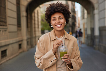 Glad curly haired woman in casual jacket drinks drinks fresh detox vegetable smoothie keeps to...
