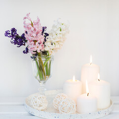 A bouquet of white, pink, blue cut hyacinth a in a small white corrugated vase and three large burning candles on a round tray are on a beige table. Place for text