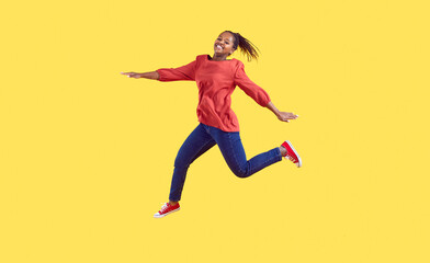 Fototapeta na wymiar Happy black woman jumping in the studio. Positive carefree young Afro American girl wearing comfortable red blouse and blue jeans jumping high in air isolated on vibrant vivid yellow colour background