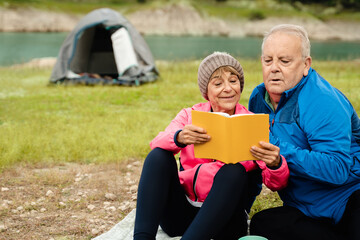 Happy senior couple having fun reading a book while camping in mountain lake - Focus on woman face