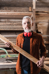 bearded woodworker with board looking at camera in workshop.