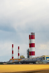 A working chemical industry plant against the background of clouds and an agricultural field. Factory pipes emit columns of smoke poisoning the environment. Poisoning of agricultural land with waste