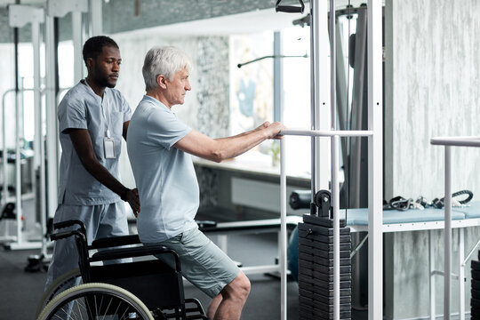 Side view portrait of senior man doing rehabilitation exercises in gym at medical clinic with therapist assisting