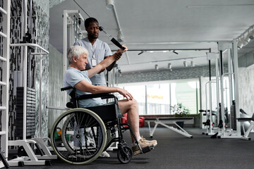 Side view portrait of senior man using wheelchair in gym and doing rehabilitation exercises with...