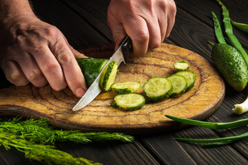Close-up of the hands of the cook with a knife cut a fresh cucumber on a wooden cutting board