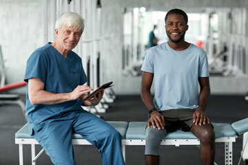Portrait of senior rehabilitation therapist smiling at camera while consulting young man in clinic