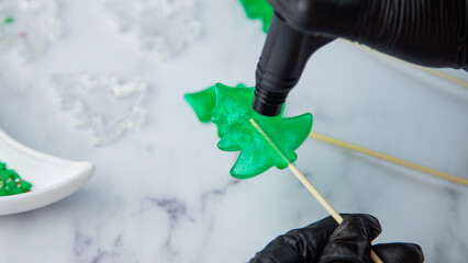 A green lollipop in the form of a Christmas tree is held in the hands of black gloves and burned on fire from a lighter, in the process of making a lollipop. High quality photo