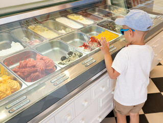 Little boy in a cap and sunglasses chooses ice cream in a cafe showcase