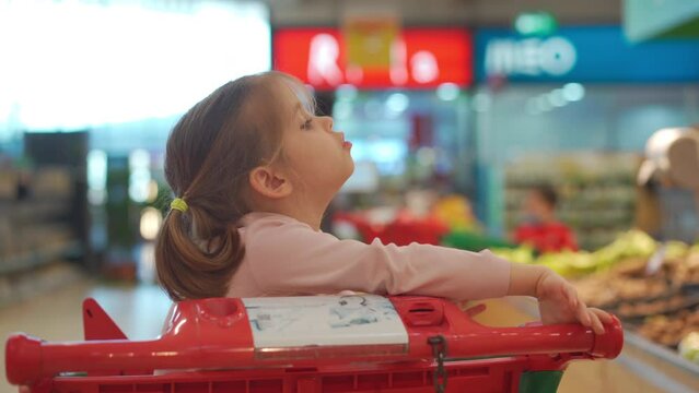Child, little girl 5 years sitting shopping cart in supermarket while mother choose goods to buy it Female kid shopping cart food store or supermarket. Little kid going shopping. Healthy food for kids