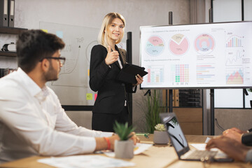 Beautiful confident caucasian businesswoman lady, showing her financial ad sales report on big digital screen showing thumb up for her focused high-skilled multiracial business colleagues.
