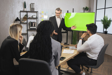 Front view of mature confident bearded businessman, showing statistic datas on green chroma key screen for his focused high-skilled multiethnical colleagues during meeting in office room.