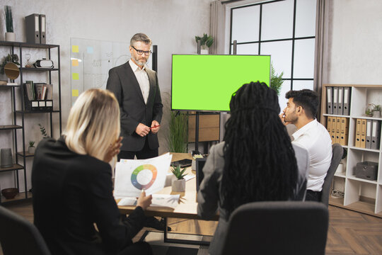 Handsome mature bearded chief, having a meeting together with his focused international colleagues, and showing business strategy on green chroma key screen with financial marketing charts.