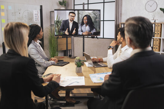 Team of diverse four multiethnic businesspeople having video meeting in boardroom at office indoor looking at African business lady and Caucasian man, using smart video technology to communicat