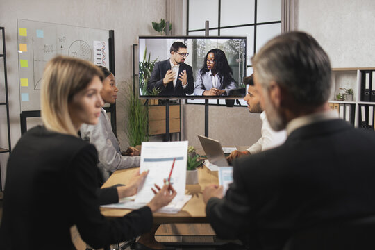 Team of diverse business people looking at screen during video conference in office, listening young pretty African woman and caucasian bisnessman, talking and gesturing. Teleconference.