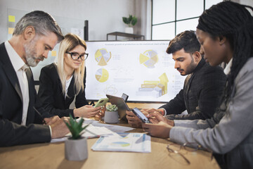 Diverse group of multiracial business people in the conference room with big TV screen, cooperating at the table, looking at papers with financial charts and statistics. Teamwork concept.