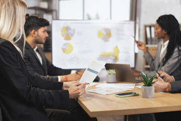 Close up cropped shot of diverse business team, having meeting in boardroom at office, sitting at the table, making notes, using laptop, tablets pc, and papers with infographics.