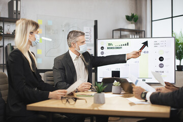 Mature bearded chief in face mask, having a meeting together with his focused international colleagues, and showing business strategy on interactive digital screen with financial marketing charts.