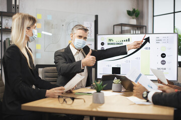 Mature bearded chief in face mask, having a meeting together with his focused international colleagues, and showing business strategy on interactive digital screen with financial marketing charts.