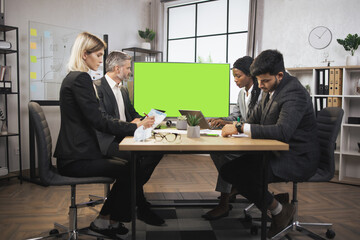 Diverse group of successful multiracial focused businesspeople in the conference room, looking at digital wall screen with green chroma key copy space. Business meeting concept.