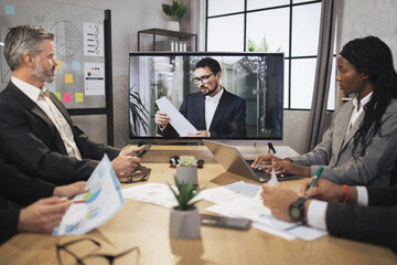 Group of four skillful multiethnical businesspeople looking attentively at screen, having video conference in boardroom, with their male colleague explaining some financial charts.