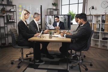 Team of four focused multiethnical businesspeople looking at screen, having video conference in modern office, with their male colleague explaining some financial charts. Telemeeting.