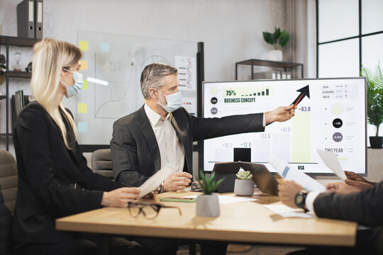 Mature bearded chief in face mask, having a meeting together with his focused international colleagues, and showing business strategy on interactive digital screen with financial marketing charts.