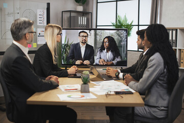 Confident Caucasian business man and African woman tells business program concept to their multinational colleagues during a video conference in a modern office with a huge TV for conversations