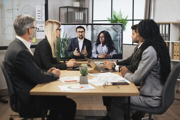 Confident Caucasian business man and African woman tells business program concept to their multinational colleagues during a video conference in a modern office with a huge TV for conversations