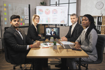 Portrait of group of four skillful multiethnical businesspeople looking at the camera while having video conference from laptop in boardroom, with their male colleague explaining some financial charts