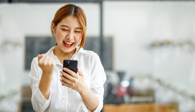 Image of a happy excited Asian young woman using a mobile phone and looking at the screen, isolated over blue wall background make winner gesture.